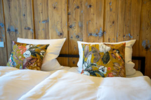 Ein ordentlich gemachtes Bett mit weißer Bettwäsche und zwei Kissen mit bunten Blumenmustern vor einer rustikalen Holzwand.
