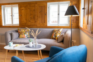Ein gemütliches Wohnzimmer in einer Ferienwohnung in der Oberlausitz mit einem grauen Sofa, das mit geblümten Kissen dekoriert ist, einem kleinen weißen Couchtisch mit einer Tasse und Vasen, einem blauen Sessel und einer schwarzen Stehlampe neben Fenstern mit Holzrahmen.
