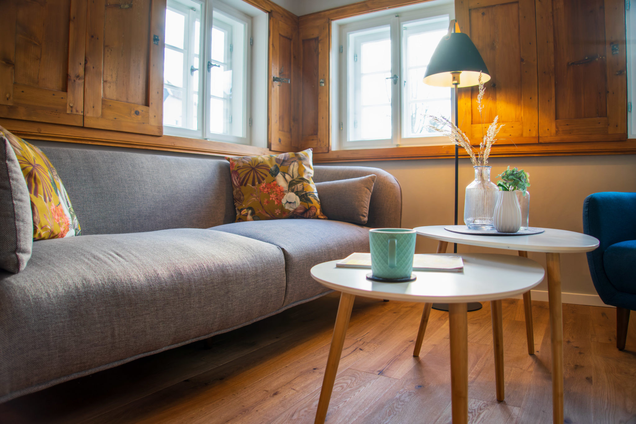 Ein gemütliches Wohnzimmer mit einem grauen Sofa, geblümten Kissen, zwei runden weißen Couchtischen, einer grünen Tasse, einem Buch, dekorativen Vasen, einer Stehlampe, holzgetäfelten Wänden und Fenstern, die natürliches Licht hereinlassen.