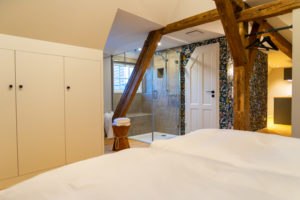 Eine moderne Ferienwohnung für zwei in der Oberlausitz mit einer gläsernen Dusche, Holzbalken, weißen Einbauschränken, einem Korb mit Handtüchern und floralen Tapetenakzenten im Badbereich.