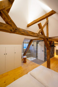 Ein gemütliches Schlafzimmer im Dachgeschoss mit sichtbaren Holzbalken, hellen Holzböden und einem weißen Bett - perfekt für Paare, die einen erholsamen Urlaub oder einen komfortablen Rückzugsort zum Arbeiten suchen. Warme Beleuchtung und ein modernes Bad schaffen eine einladende Atmosphäre.