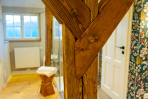 Nahaufnahme eines Holzbalkens mit sichtbaren Fugen in einem hellen, modernen Badezimmer mit Blumentapete, weißer Tür, Holzfußboden und einem Handtuch auf einem Holzhocker neben einer Glasdusche.