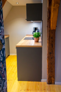 Eine moderne Küche mit hölzernen Arbeitsplatten, grauen Schränken, einer Basilikum-Topfpflanze, einer Kaffeemaschine und einem Wasserkocher. Der Fußboden ist aus hellem Holz, und an der Wand befinden sich freiliegende Holzbalken.