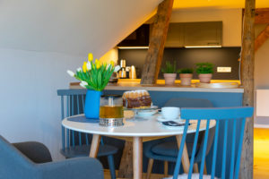 Eine gemütliche Küche in einer Ferienwohnung für zwei in der Oberlausitz mit Holzbalken, einem kleinen runden Tisch für Tee und Kuchen, gelben und weißen Tulpen in einer Vase und Topfkräutern auf dem Tresen im Hintergrund.