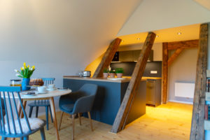 Moderne Dachbodenküche und Essbereich mit sichtbaren Holzbalken, hellen Holzböden, einem kleinen Frühstückstisch, blauen Stühlen und Topfpflanzen auf dem Tresen. Helle, gemütliche und einladende Atmosphäre der Ferienwohnung Carl in Neusalza-Spremberg.