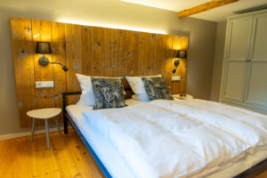 Ein gemütliches Schlafzimmer in einer Ferienwohnung in Sachsen mit einem Doppelbett, weißer Bettwäsche, blattgemusterten Kissen, einem hölzernen Kopfteil mit eingebauten Leselampen, einem kleinen weißen Beistelltisch, einem Kleiderschrank und einem Holzboden - perfekt für Urlaub in Deutschland.