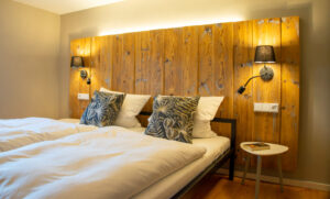 Zwei sauber gemachte Einzelbetten mit weißem Bettzeug und gemusterten Kissen stehen nebeneinander. Hinter ihnen steht ein rustikales Holzkopfteil mit zwei Wandlampen, und neben den Betten steht ein kleiner runder Tisch. Warmes, gemütliches Licht erfüllt den Raum.