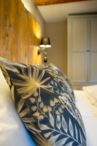 Nahaufnahme eines Bettes mit dekorativen Kissen mit botanischem Muster in neutralen Tönen, einem Kopfteil aus Holz und einer Wandlampe neben einem Kleiderschrank in einem gemütlichen, sanft beleuchteten Schlafzimmer.