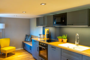 Moderne Küche mit grauen Schränken, hölzernen Arbeitsplatten, einem eingebauten Ofen, einer Mikrowelle, einem Spülbecken, Topfpflanzen und Küchenzubehör. Auf der linken Seite befindet sich eine gemütliche Sitzecke mit blauen Kissen und einem gelben Sessel.