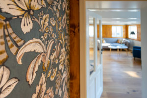 Nahaufnahme einer Wand mit Tapete mit Blumenmuster neben einer offenen Tür, die in einen hellen Raum mit Holzböden, modernen Möbeln und gesprossten Fenstern führt, die natürliches Licht hereinlassen. Wir blicken vom Flur in die historische Blockstube der Ferienwohnung Alwine im MARKTQUARTIER 5.