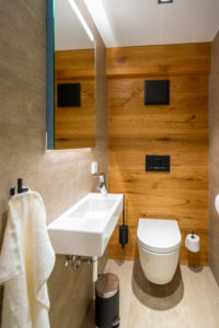 Ein modernes Badezimmer mit Holzvertäfelung, beigen Fliesenwänden, einer wandmontierten Toilette, einem rechteckigen Waschbecken mit Handtuch- und Toilettenpapierhalter und einem großen Spiegel über dem Waschbecken. Der Raum ist gut beleuchtet und minimalistisch.