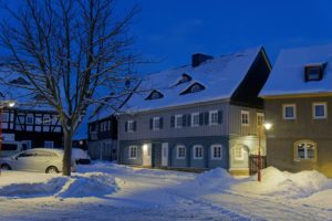 Eine verschneite Straßenszene in der Abenddämmerung zeigt traditionelle Häuser mit schneebedeckten Dächern, einen blattlosen Baum und ein geparktes Auto. Eine Straßenlaterne wirft einen warmen Schein auf die winterliche Landschaft. Gemütlicher Winterurlaub in der Oberlausitz.
