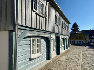 Ein traditionelles Holzhaus mit blaugrau gestrichenen Wänden und weiß gerahmten Fenstern steht in einer Kopfsteinpflasterstraße unter einem klaren blauen Himmel, im Hintergrund sind weitere bunte Häuser zu sehen.