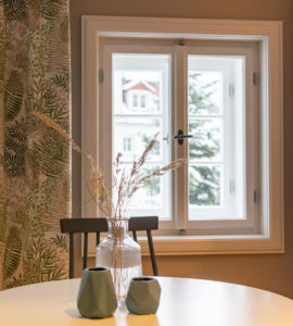 Ein runder weißer Tisch mit einer Glasvase mit getrockneten Gräsern und zwei grünen geometrischen Vasen vor einem Fenster mit Blick auf Häuser und Bäume; links hängen gemusterte Vorhänge.