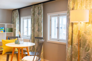 Ein gemütliches Kinderzimmer in der Ferienwohnung mit einem runden weißen Tisch, zwei grauen Stühlen, grün gemusterten Vorhängen, einer hohen Stehlampe und Regalen mit Spielen und Büchern neben zwei Fenstern.