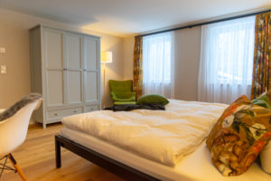 Ein gemütliches Schlafzimmer mit einem ordentlich gemachten Bett, bunten Kissen mit Blumenmuster, einem grauen Kleiderschrank, einem olivgrünen Sessel, einer Stehlampe, hellem Holzboden und großen Fenstern mit durchsichtigen weißen Vorhängen und gemusterten Gardinen.