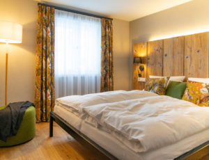 Ein gemütliches Schlafzimmer in der Ferienwohnung Alwine im Marktquartier 5 verfügt über ein großes Bett mit geblümten Kissen und bunten Vorhängen. Ein grüner Stuhl mit einer dunklen Decke sitzt neben einer hohen Lampe, während das Sonnenlicht durch die weißen Vorhänge strömt und für ein wirklich gemütliches Gefühl sorgt.