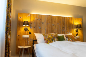 Gemütliches Schlafzimmer in einer Ferienwohnung in Neusalza-Spremberg mit einem großen Holzkopfteil, schwarzen Wandlampen, weißer Bettwäsche und bunten Kissen. Ein runder Nachttisch mit einem Buch steht neben dem Bett, während von links natürliches Licht einfällt.