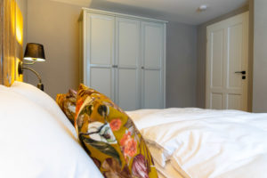 Ein gemütliches Schlafzimmer im Marktquartier 5 in der Ferienwohnung in der Oberlausitz ist mit einem ordentlich gemachten Bett, geblümten Kissen, einer schwarzen Nachttischlampe und einem großen weißen Schrank vor einer hellen Wand ausgestattet.