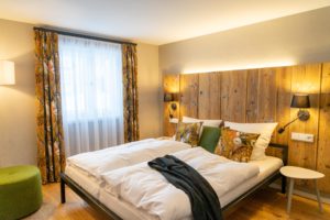 Ein gemütliches, helles Schlafzimmer in einer Ferienwohnung in der Oberlausitz mit einem Doppelbett mit bunten, geblümten Kissen, einem Kopfteil aus Holz, Nachttischlampen, geblümten Vorhängen an einem großen Fenster, einem grünen Hocker und einem weißen Beistelltisch.