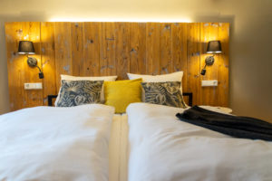 Ein gemütliches Bett mit weißer Bettwäsche, dekorativen Kissen, darunter ein gelbes Akzentkissen, ein rustikales Kopfteil aus Holz und zwei Wandlampen mit schwarzen Lampenschirmen auf beiden Seiten.