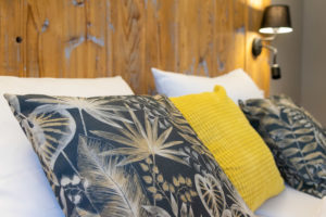 Nahaufnahme eines Bettes mit weißen Kissen, zwei dunkelblauen und weißen blattgemusterten Kissen und einem gelben strukturierten Kissen vor einem rustikalen Holzkopfteil und einer Wandlampe.