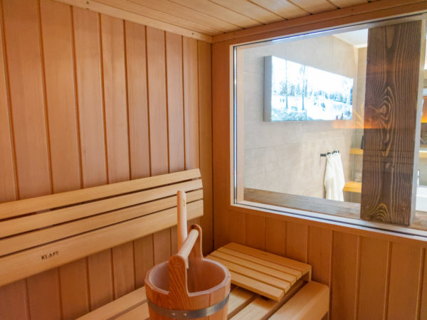 Eine Holzsauna mit Bänken und einem Holzkübel. Durch ein großes Glasfenster sieht man ein Badezimmer mit einer Badewanne und einem montierten TV-Bildschirm.