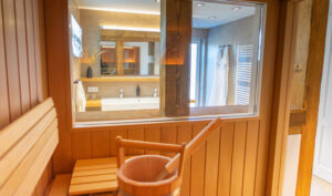 Blick aus dem Inneren einer Holzsauna durch ein Glasfenster in ein modernes Badezimmer mit Waschbecken, Spiegel, Handtuchhalter und einem hängenden Bademantel. Auf der Saunabank im Vordergrund stehen ein hölzerner Eimer und eine Schöpfkelle.