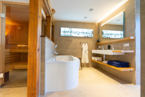 Modernes Badezimmer mit freistehender Badewanne, Doppelwaschbecken, Holzregalen, zwei Spiegeln, Wandkunst und einem Blick in die Saunakabine links. Handtücher, Bademäntel und warmes Licht sorgen für eine gemütliche Atmosphäre.
