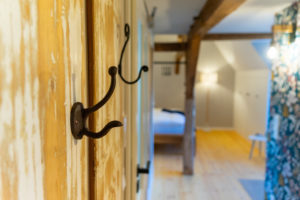 Nahaufnahme eines metallenen Wandhakens an einer holzgetäfelten Wand in einem hellen, rustikalen Raum mit Holzböden, freiliegenden Balken und unscharfer Ansicht eines Bettes und Mobiliars im Hintergrund.