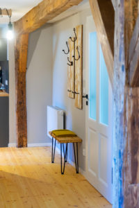 Ein Flur mit Holzbalken, hellem Holzboden, einer weißen Tür, Wandgarderobenständern und einer kleinen Bank mit zwei gelben Kissen unter den Ständern. Links ist eine hängende Glühbirnenlampe zu sehen.