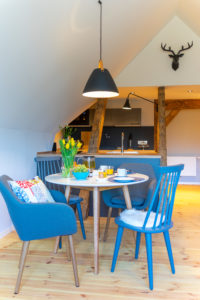 Eine gemütliche, moderne Küche und ein Essbereich mit blauen Stühlen um einen kleinen runden Tisch, der für das Frühstück gedeckt ist. Gelbe Tulpen, Obst und Saft stehen auf dem Tisch; Holzbalken und ein Hirschkopf an der Wand zieren den Hintergrund. Ein perfekter Urlaubsmorgen im Marktquartier 5.