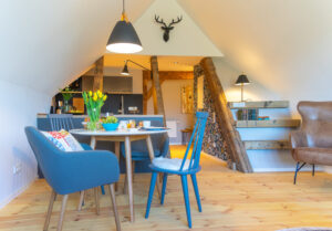 Eine gemütliche Essecke im Dachgeschoss der Ferienwohnung in Neusalza-Spremberg, mit einem runden Holztisch für das Frühstück, blauen und grauen Stühlen, frischen Tulpen, rustikalen Balken, moderner Beleuchtung und einem dekorativen Hirschkopf über der Küche.