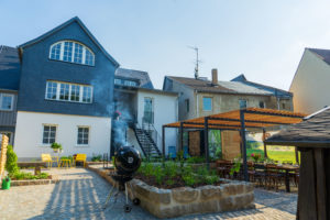 Ein geräumiger Garten, ideal für Familienurlaub oder Familientreffen, mit einem Grill zum Rauchen, Hochbeeten, einem Esstisch mit Stühlen und einer Pergola neben einem großen, mehrstöckigen Haus mit einer grau-weißen Fassade.