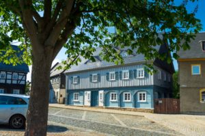 Ein großer, belaubter Baum spendet Schatten auf einem gepflasterten Platz vor einem traditionellen blau-weißen Fachwerkhaus – perfekt für einen erholsamen Aufenthalt in einer Ferienwohnung in der Oberlausitz – vor einem klaren Himmel, mit einem silbernen Auto, das links geparkt ist.
