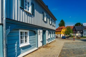 Eine Kopfsteinpflasterstraße schlängelt sich an einem blauen Holzhaus mit weiß gerahmten Fenstern vorbei. Im Hintergrund sind bunte Häuser und einige Bäume unter einem klaren blauen Himmel zu sehen.