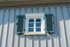 Ein kleines weißgerahmtes Fenster mit blauen Fensterläden befindet sich in einer hellgrauen Holzwand, typisch für charmante Ferienwohnungen in Sachsen. Das Fenster gehört zum Marktquartier 5, dem perfekten Ort für Urlaub im Denkmal. Sonnenlicht wirft dekorative Schatten auf die vertikalen Paneele und Zierleisten.