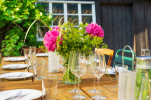 Ein hölzerner Esstisch im Freien ist mit Gläsern, Tellern und einem Mittelstück aus rosa Blumen und Grünzeug gedeckt. Helles Sonnenlicht schafft eine fröhliche, von der Landlust inspirierte Atmosphäre, die perfekt für Urlaubsmomente ist. Urlaub in der Oberlausitz.