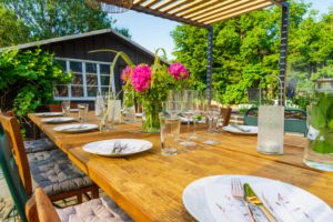 Ein hölzerner Esstisch im Freien, gedeckt für eine Mahlzeit mit Tellern, Besteck, Gläsern und einer Vase mit rosa Blumen, unter einer Pergola mit Grünzeug und einem Gartenhaus im Hintergrund an einem sonnigen Tag.