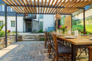 Ein langer Holztisch mit Stühlen steht unter einer Pergola im sonnigen Innenhof von Marktquartier 5 - perfekt für ein Familientreffen oder einen Urlaub. Der Tisch ist mit Gläsern und Blumen gedeckt, mit einem modernen weißen Haus und einer Wendeltreppe im Hintergrund.