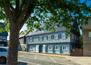 Ein großer Baum spendet an einem sonnigen Tag Schatten auf dem Kopfsteinpflaster vor einem traditionellen blau-weißen Fachwerkhaus mit Fensterläden und Dachgauben – ein einladender Anblick auf ein Ferienhaus in der Oberlausitz. Auf der Straße ist ein geparktes Auto zu sehen.