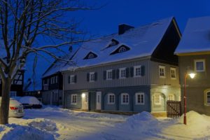 Ein traditionelles Haus mit schneebedecktem Dach und schneebedeckten Fenstern leuchtet in der Dämmerung warm. Schnee türmt sich rund um das Gebäude und parkende Autos in einer ruhigen, verschneiten Straße unter einem tiefblauen Abendhimmel.