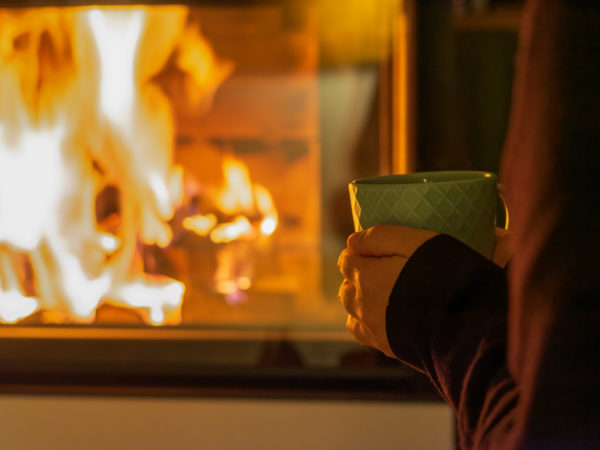 Eine Person mit einer grünen Tasse steht neben einem warmen Kamin, hinter dessen Glasscheibe orangefarbene Flammen glühen und eine gemütliche Atmosphäre schaffen.