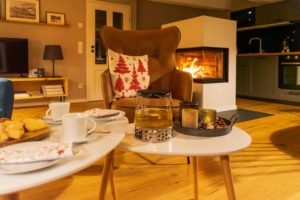 Ein gemütliches Wohnzimmer mit einem braunen Sessel, einem festlichen Kissen, einem brennenden Kamin und einer Küche im Hintergrund. Auf dem weißen Couchtisch stehen Tee, Tassen, Kekse, Kerzen und weihnachtliche Servietten.