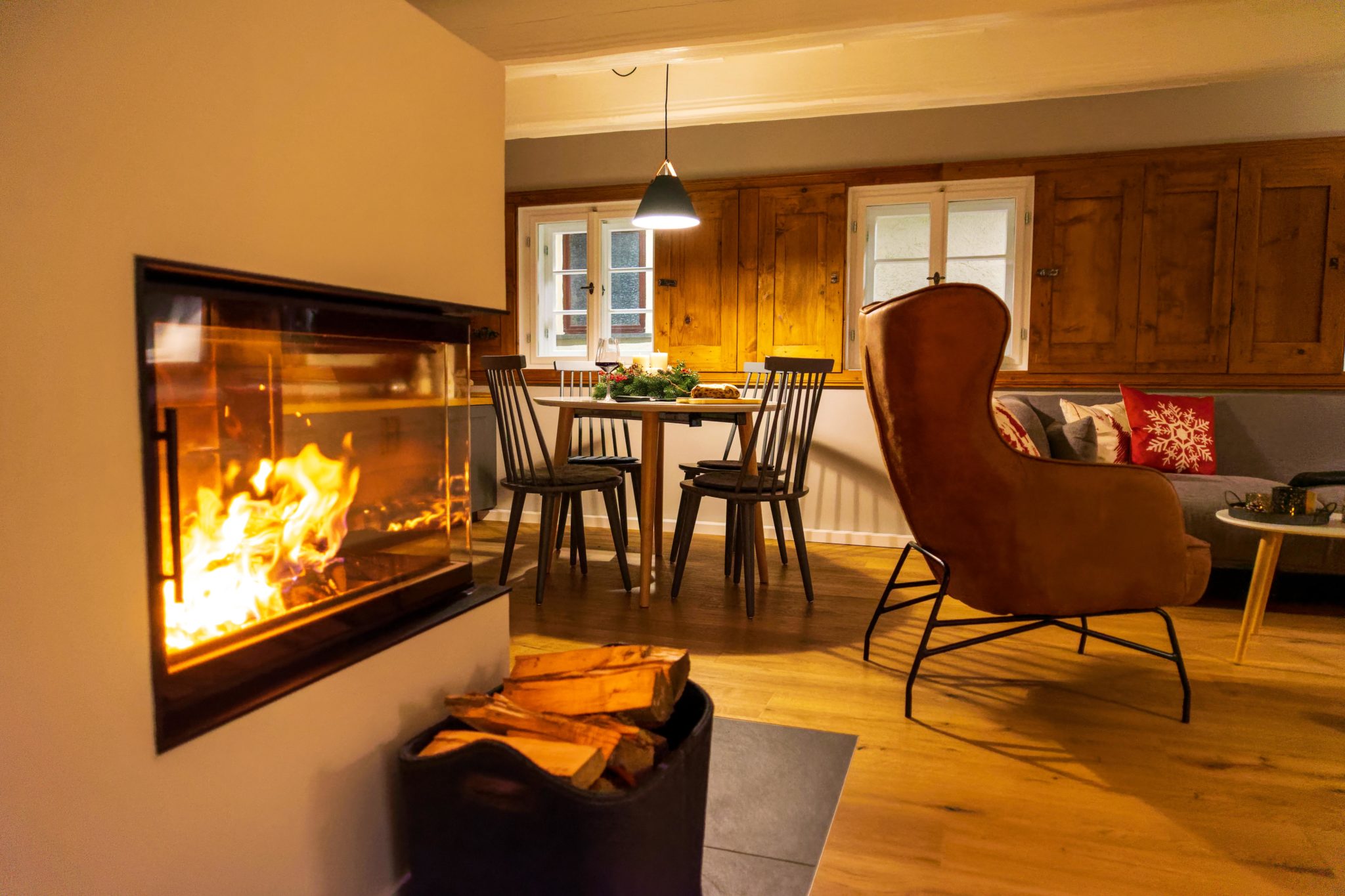 Ein gemütliches Wohnzimmer mit brennendem Kamin, einem Stapel Brennholz, einem braunen Sessel, einem kleinen runden Esstisch mit Stühlen und warmer Beleuchtung, die eine einladende Atmosphäre schafft.