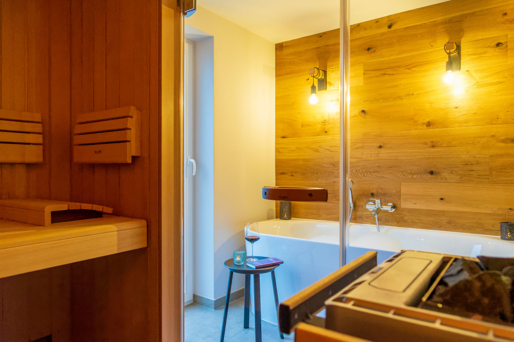 Modernes Badezimmer mit Holzsauna links, weißer Badewanne, Holzpaneelen, zwei Wandleuchten und einem kleinen Tisch neben der Wanne mit einem Getränk und einem Buch. Warmes Licht sorgt für eine gemütliche Atmosphäre.