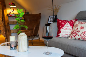 Ein gemütliches, weihnachtlich dekoriertes Wohnzimmer mit festlichen Kissen auf einem grauen Sofa, einem Glas Rotwein auf einem weißen Couchtisch, einer Holzvase mit Stechpalme und einer Kerze. Im Hintergrund sind eine warme Beleuchtung und ein brauner Sessel zu sehen.