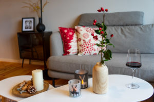 In einem gemütlichen Wohnzimmer einer Ferienwohnung sorgen ein graues Sofa mit roten und weißen Weihnachtskissen, ein Tisch mit einer Vase mit roten Beeren, Kerzen, ein Glas Rotwein und festliche Dekoration für eine warme und einladende Atmosphäre.