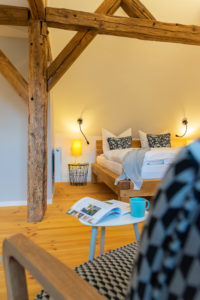 Gemütliches Schlafzimmer im Ferienapartment Carl im Ferienhaus Marktquartier 5 in Neusalza-Spremberg mit freiliegenden Holzbalken, einem Bett mit weißer Bettwäsche und gemusterten Kissen, einem kleinen Tisch mit einer aufgeschlagenen Zeitschrift und einer blauen Tasse sowie einer warmen Beleuchtung, die eine einladende Atmosphäre schafft.