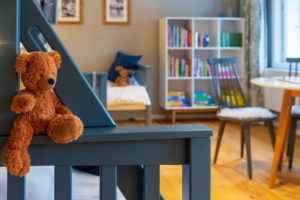 Ein brauner Teddybär sitzt auf dem Geländer eines blauen Bettes im gemütlichen, hellen Kinderschlafzimmer in der Ferienwohnung Hermann im Marktquartier 5, mit Bücherregalen, Stühlen und gerahmten Bildern im Hintergrund.